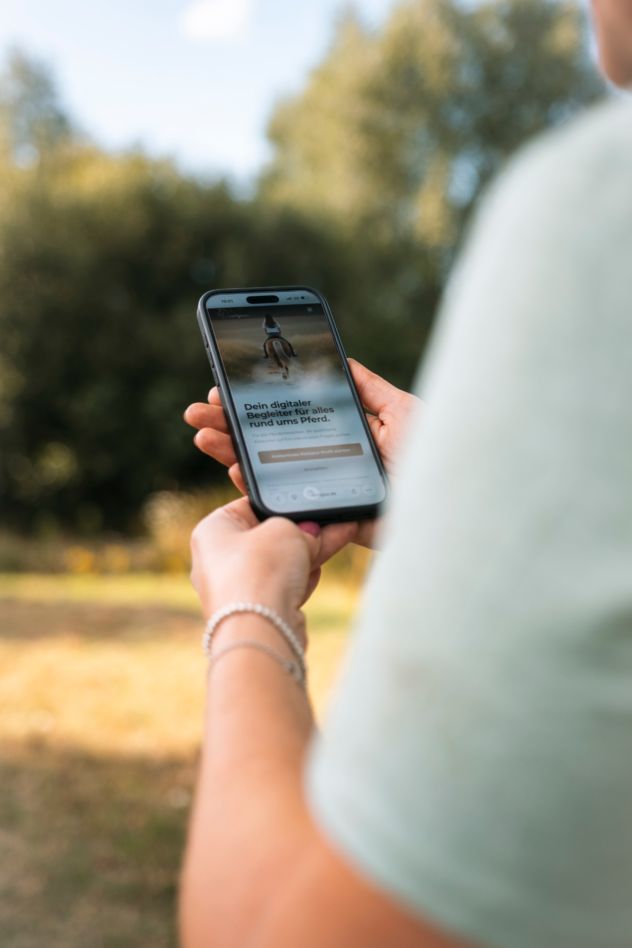 Reiterin hält ein Smartphone mit der geöffneten Reitspur-App, dahinter Bäume und Wiese im Abendlicht